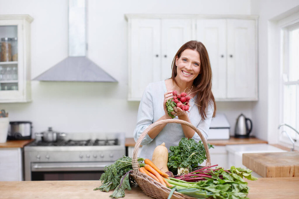 Rábanos encantadores. Una mujer hermosa sosteniendo un montón de rábanos mientras está de pie detrás de una cesta de verduras frescas en su cocina - Foto, Imagen