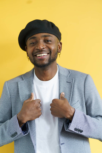Happy stylish african american man smiling isolated on yellow background. Young student with earrings in suit and beret posing for camera. High quality photo - Foto, immagini