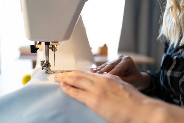 Zamknij widok na proces szycia. Kobiece ręce szyjące niebieską tkaninę na profesjonalnej maszynie do szycia w miejscu pracy. zamazane tło. Przestrzeń kopiowania - Zdjęcie, obraz