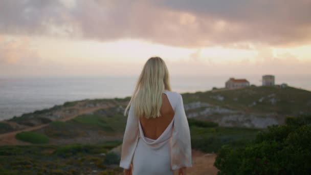 Flirtująca blondynka spacerująca zamkowy krajobraz zbliżenie. Zmysłowa kobieta patrząca przez ramię na zachody słońca, widok z tyłu. Wyrafinowany model w sukience otwartej opartej na wieczornym brzegu morza - Materiał filmowy, wideo