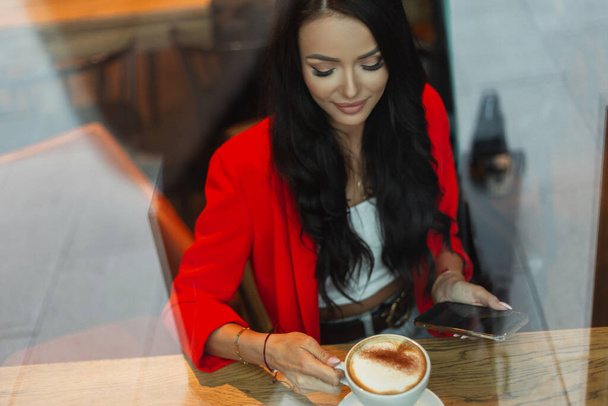 Hermosa empresaria exitosa en ropa brillante de moda con una chaqueta roja está sentado en un café con un teléfono inteligente, beber café. Chica descanso en la cafetería detrás de vidrio con reflexión - Foto, Imagen