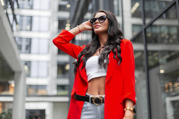 Mooie succesvolle modieuze zakenvrouw met krullend haar en modezonnebril met een mode rode blazer, top, jeans en een portemonnee wandelingen in de stad in de buurt van moderne gebouwen - Foto, afbeelding