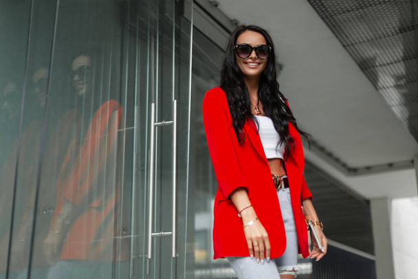 Felicidad joven hermosa mujer elegante con sonrisa con gafas de sol de moda en traje de negocios casual de moda con chaqueta roja, top y jeans camina por la calle en un fondo urbano moderno - Foto, imagen