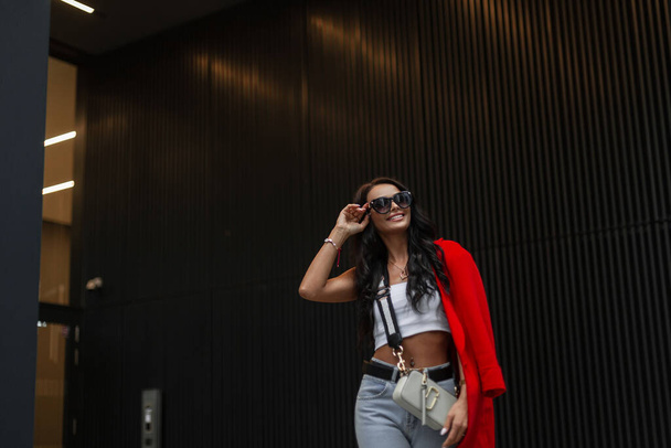 Muoti kaunis liiketoiminnan tyttö tyylikäs rento vaatteita alkuun, farkut, punainen takki ja laukku kävely kaupungissa ja yllään tyylikäs aurinkolasit lähellä modernia rakennusta - Valokuva, kuva