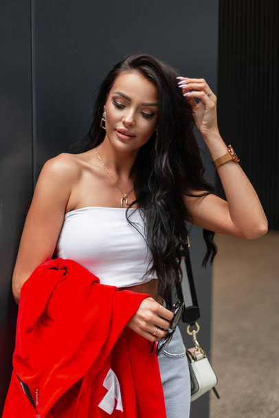 Belle dame d'affaires à la mode dans des vêtements décontractés élégants avec un haut et un jean tient veste rouge près d'un bâtiment moderne sombre dans la rue. - Photo, image