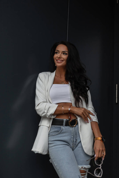 Donna di moda di bellezza urbana in eleganti abiti da lavoro con top bianco, blazer e jeans strappati con borsa di moda vicino a una parete di metallo nero  - Foto, immagini