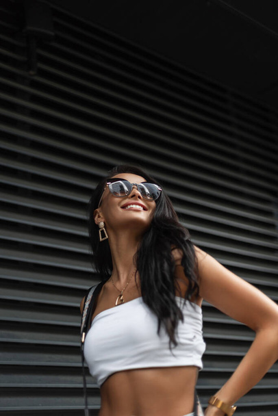 Retrato de mujer urbana feliz de una hermosa chica de moda con una sonrisa con gafas de sol en un top blanco de moda con una bolsa camina por la calle cerca de una pared de metal negro - Foto, imagen