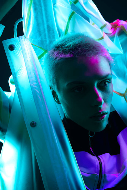 Young woman with short hair pulling up hood of futuristic coat and looking away under colorful neon illumination - Foto, Imagem