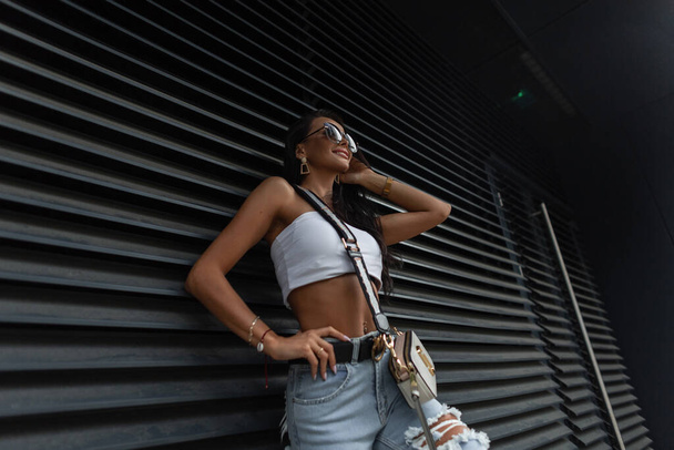 Módní krásná stylová žena model s úsměvem s módními slunečními brýlemi v létě trendy oblečení s top, roztržené džíny a malý sáček v blízkosti černé kovové zdi ve městě - Fotografie, Obrázek