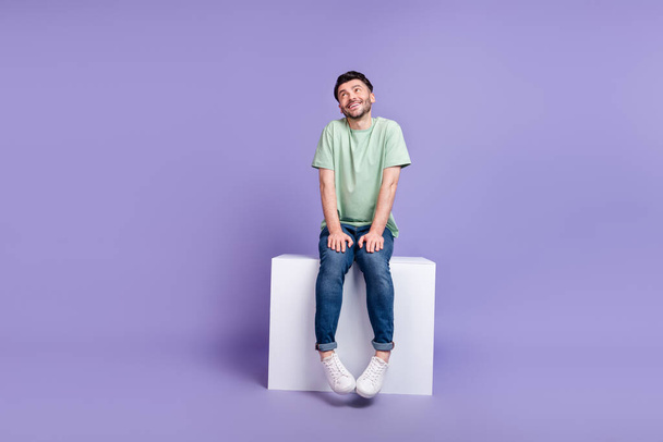 Full size photo of handsome young guy sit shy pose white cube look empty space wear trendy gray outfit isolated on violet color background. - Foto, imagen