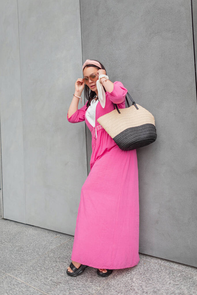 Modieus mooi meisje plus size met een bandana in stijlvolle zomerkleding met een roze pareo en een rieten strozak staat naast een grijze betonnen muur en zet een zonnebril op - Foto, afbeelding