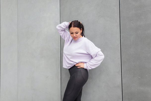 Schöne modische Body Positive Frau in stylischer Sportbekleidung mit schwarzen Leggings und einem lila Sweatshirt in Übergröße posiert in der Nähe einer grauen Betonwand - Foto, Bild