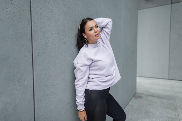 Mujer bonita de moda más modelo de tamaño en traje urbano deportivo con púrpura elegante jersey de gran tamaño y polainas negras se encuentra cerca de una pared gris de hormigón en la calle - Foto, imagen