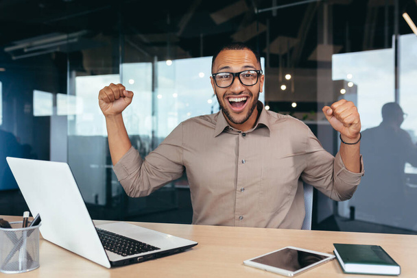 Happy and successful businessman celebrating achievement, portrait of man at work in office looking at camera and holding hands up, celebrating victory and triumph, employee using laptop. - Foto, afbeelding