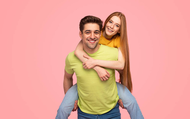 Happy young man giving piggyback ride to redhead girlfriend and looking at camera against pink background - Foto, Imagen