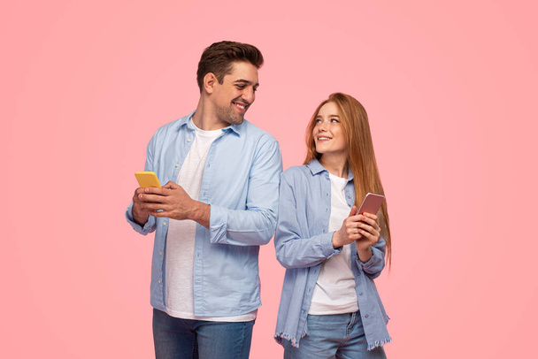 Young boyfriend and girlfriend in similar clothes browsing social media on cellphones and looking at each other with smile against pink background - Foto, afbeelding
