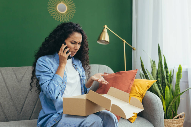 Nervous young beautiful Latin American woman is begging on the phone at home on the sofa, holding a bag, a box that was sent to her by mistake. Solves the delivery problem, of damaged goods. - 写真・画像