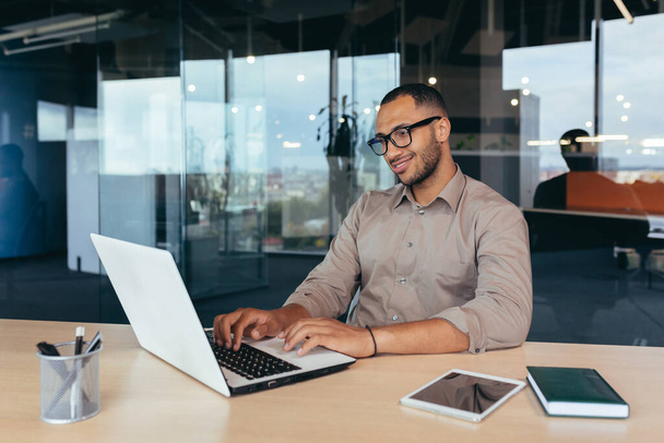 Αφροαμερικανός προγραμματιστής στην εργασία με το lap-top στο εσωτερικό γραφείο, ο άνθρωπος με τα γυαλιά και casual κώδικα γραφής για τα προγράμματα. - Φωτογραφία, εικόνα