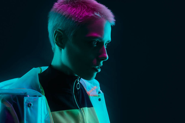 Androgynous young woman in futuristic raincoat and sweatshirt looking away while standing under colorful neon light against black background - Photo, Image