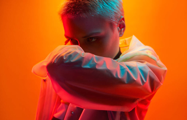 Young androgynous female model in futuristic raincoat covering face with bent arm and looking away under neon illumination against orange background - Foto, immagini