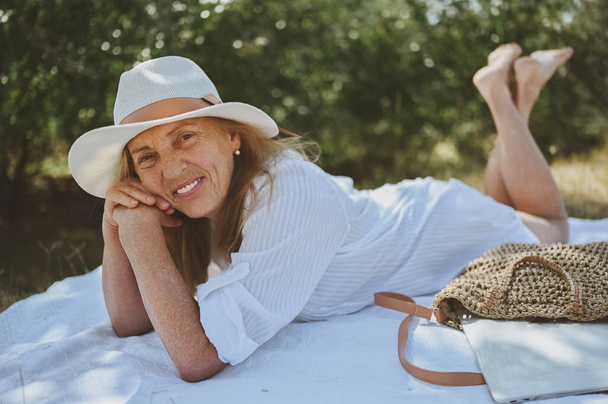 Senior happy woman laying on picnic blanket with laptop in bag at summer outdoors. Old elegant lady in straw hat on grass at countryside. Active retired people concept - Foto, Imagem