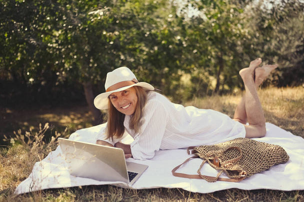 Senior happy woman working or having video call with laptop in summer outdoors. Old elegant lady in straw hat and bag sitting on grass during picnic at countryside. Active retired people concept - Foto, Bild