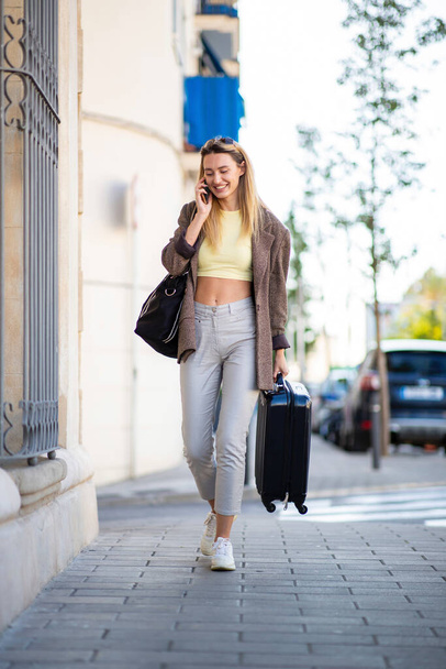 Beautiful young woman talking on mobile phone while carrying suitcase outdoors in city - Foto, Bild