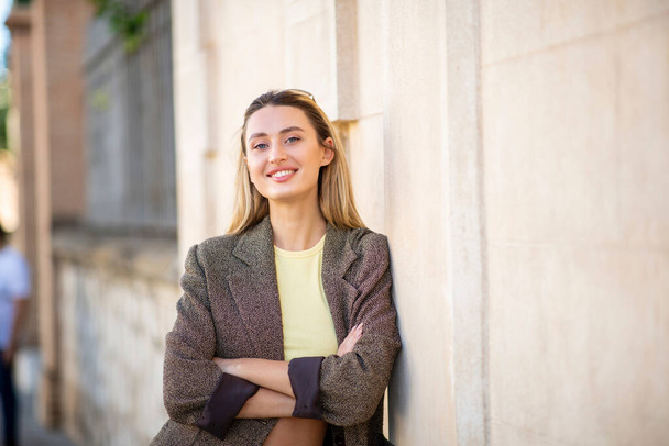 Portrait of happy stylish woman leaning against exterior wall with arms crossed - Photo, Image