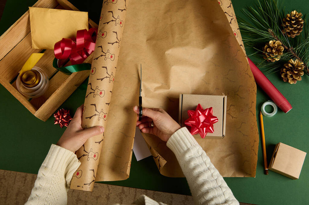 Top view woman in warm beige sweater, using scissors, cuts a wrapping paper with deer pattern, for packing gift for Christmas, New Year or other celebration event. Boxing Day. Diy present. Copy space - Photo, image