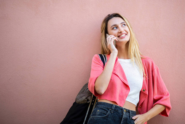 Портрет улыбающейся блондинки, разговаривающей с телефоном на розовом фоне  - Фото, изображение