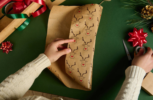 Top view of woman's hands wrapping a present for Christmas or New Year's events, using beautiful decorative paper with deer pattern, over green background. Boxing Day. Xmas preparations. Copy ad space - Photo, image