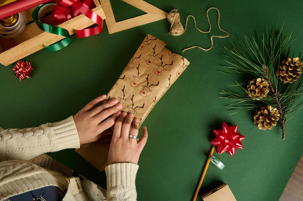 Detailní záběr. Top pohled žena balení dárek v dekorativním papíru s jelení vzor, přes zelené pozadí s obalovými materiály a zlaté borovice kužely jako vánoční výzdoba. Dárek od Diy. 2. svátek - Fotografie, Obrázek