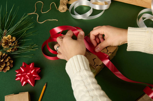 装飾的な光沢のある赤いリボンでクリスマスプレゼントを結ぶ女性の手のオーバーヘッドビューは、包装材を配置した緑の背景に。手仕事の創造的な芸術。梱包用ダイプレゼント - 写真・画像