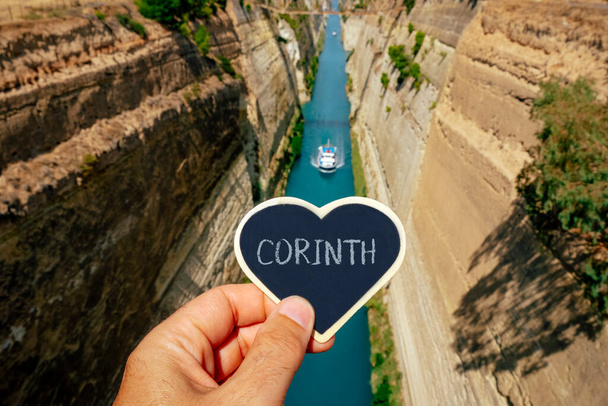 Nahaufnahme der Hand eines Mannes, der an einem Sommertag am Kanal von Korinth in Griechenland ein schwarzes herzförmiges Schild mit dem Text Korinth in der Hand hält - Foto, Bild