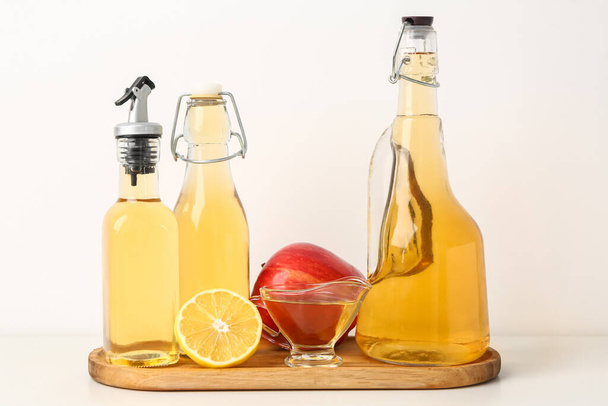 Wooden board with bottles of apple cider vinegar and fruits on light background - Фото, изображение