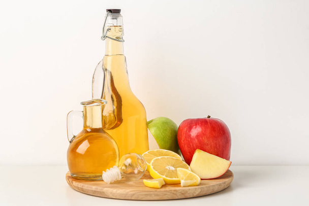 Wooden board with bottles of apple cider vinegar and fresh fruits on light background - Photo, image