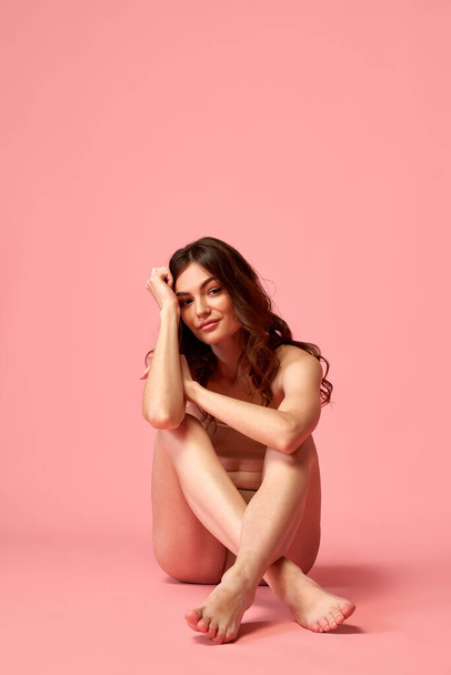 Portrait of young beautiful woman posing, sitting pon floor in beige underwear isolated over pink studio background. Femininity. Concept of beauty, fitness, spa, cosmetology, skin and body care - 写真・画像