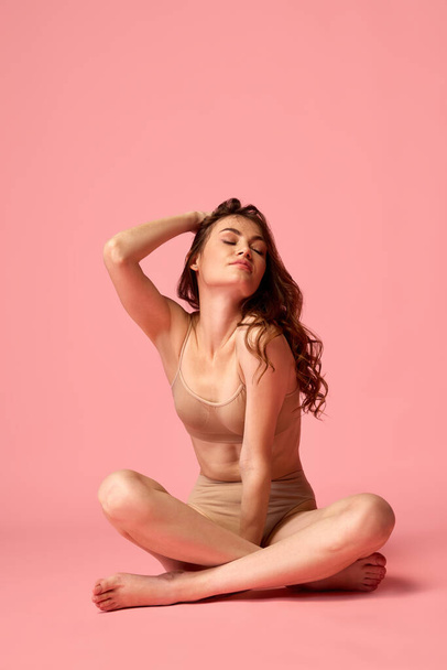 Portrait of young beautiful woman with curly hair sitting on floor, posing in beige underwear isolated over pink studio background. Concept of beauty, fitness, spa, cosmetology, skin and body care - Φωτογραφία, εικόνα