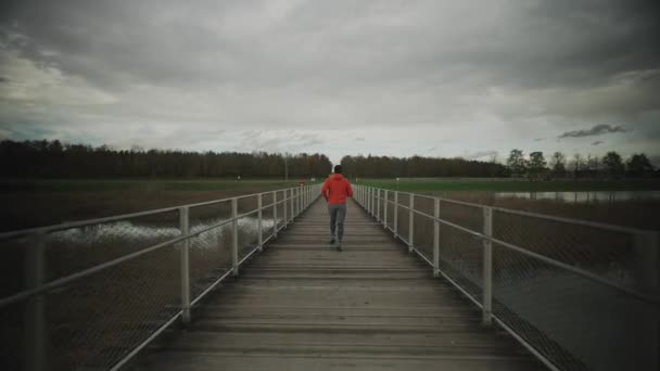 Hombre corredor en sudadera naranja corre a través de puente de madera en la naturaleza en Escandinavia en otoño. Atleta corre a través de la pasarela en tiempo frío. Estilo de vida saludable. Recorrido por el parque nacional.  - Imágenes, Vídeo