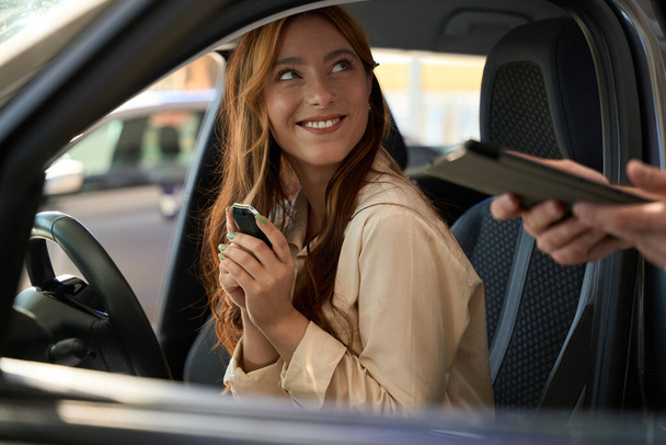 Happy lady with auto key fob sitting at steering wheel and looking at salesperson with tablet computer in hands - Photo, Image
