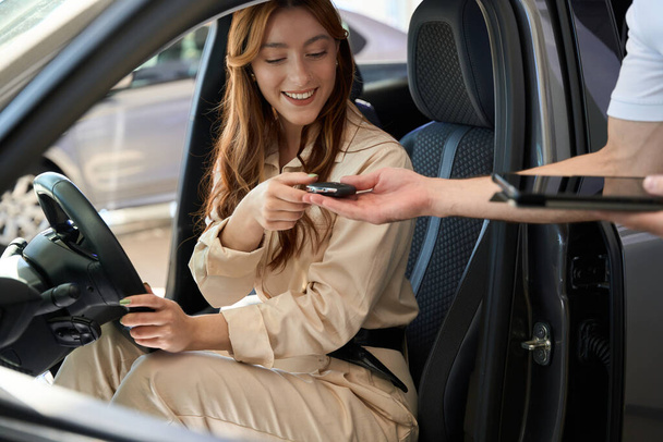 Happy vehicle buyer seated behind steering wheel getting car key fob from sales consultant - Foto, imagen
