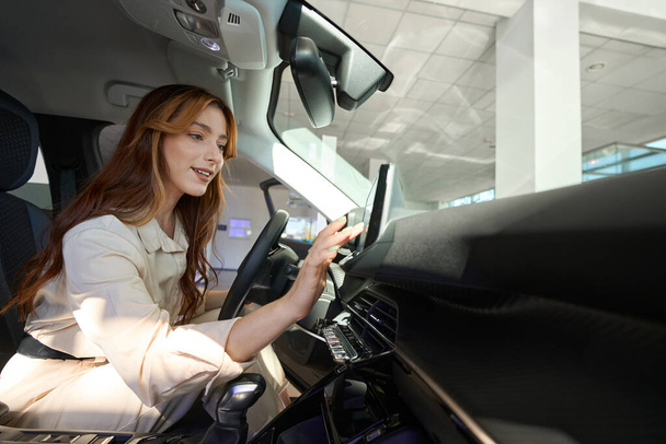 Smiling young woman seated in driver seat tapping on vehicle on-board computer - 写真・画像