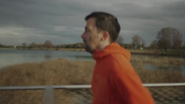 Hombre con capucha naranja corriendo por el parque nacional a través de un puente de madera en el desierto en el frío clima de otoño. Deportista hace un entrenamiento de fitness trotando en una pasarela de madera en el campo.  - Metraje, vídeo