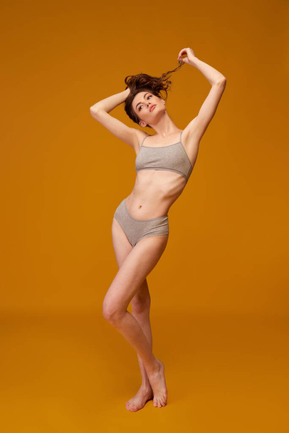 Portrait of young beautiful woman with curly hair posing in grey underwear isolated over yellow studio background. Concept of beauty, fitness, spa, cosmetology, skin and body care, diet - Φωτογραφία, εικόνα