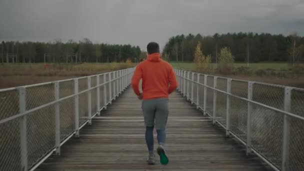 Correr en clima frío de otoño en la naturaleza. Un hombre cruza un puente de madera en el parque nacional. Un atleta en entrenamiento a campo traviesa. Entrenamiento en Canadá. Deporte activo y fitness al aire libre. Estilo de vida saludable - Imágenes, Vídeo