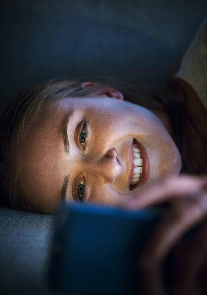 Woman, phone or lying on sofa at night in house or home living room on online dating app, social media or internet esports game. Smile, happy or relax student on mobile technology social network news. - Photo, Image