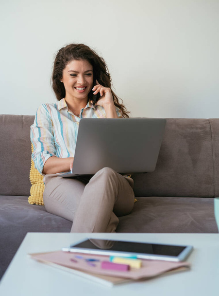 Szczęśliwa bizneswoman rozmawiająca przez telefon komórkowy i wpisująca raport biznesowy na klawiaturze laptopa siedząc na kanapie w salonie - Zdjęcie, obraz