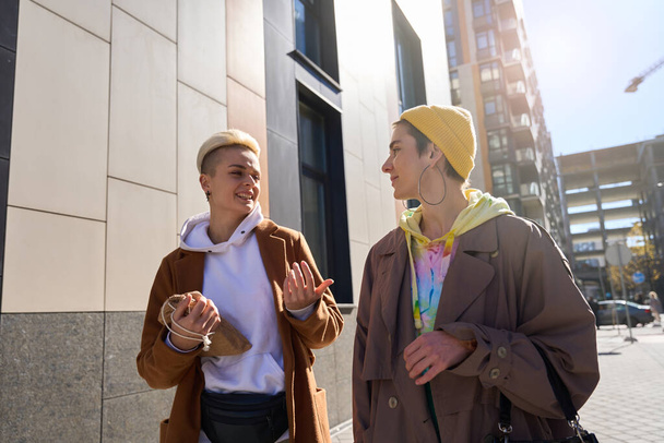 Blonde with stylish haircut and brunette in yellow cap, lgbt couple, communicate with pleasure and walk around city on day off - 写真・画像