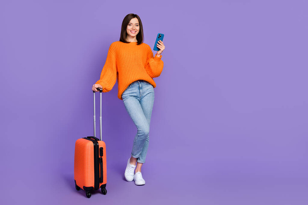 Full body photo of gorgeous young girl hold gadget booking hotel dressed stylish orange knitted look isolated on purple color background. - Foto, immagini