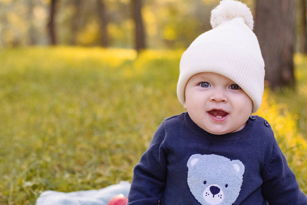 dítě v pleteném klobouku sedí na trávě v parku na pozadí podzimních stromů. dětské oblečení. venčit venku. Kvalitní fotografie - Fotografie, Obrázek
